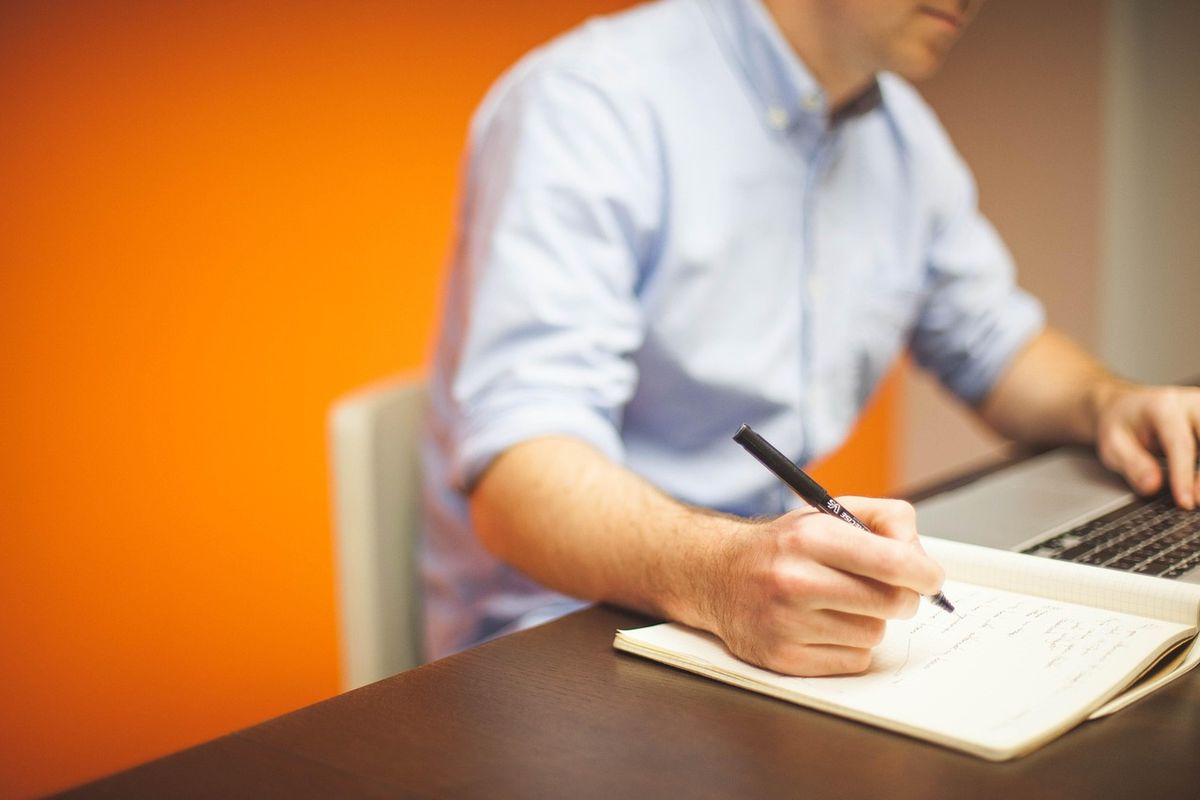 Man in a blue shirt writing notes beside his laptop