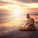 Monk in orange robes meditating on a beach at sunset