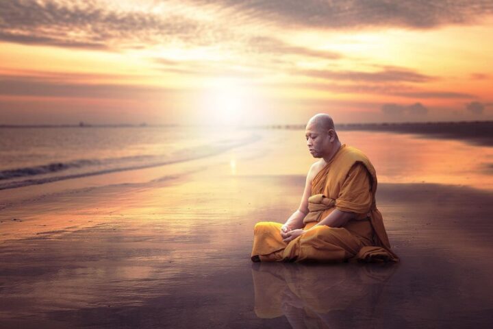 Monk in orange robes meditating on a beach at sunset