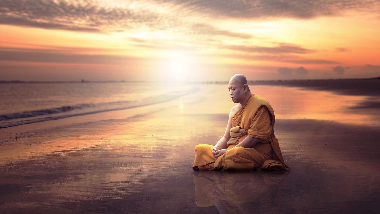 Monk in orange robes meditating on a beach at sunset