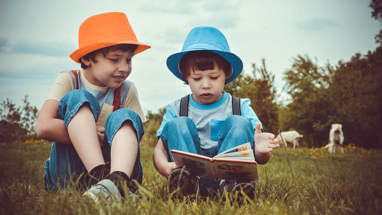 Two boys in colourful hats reading a book on the grass